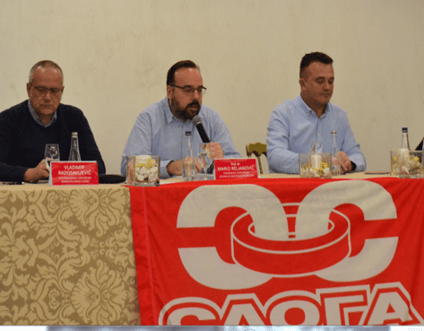 Тројица мушкараца за столом са микрофоном и банером Слога